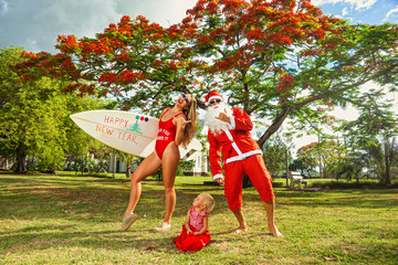 A young family of surfers and baby in Santa's costumes celebrate Christmas on a tropical island