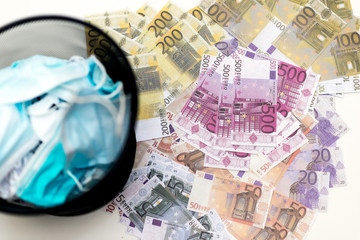 Top view close up medical protective face masks are in black trash bin on background of lots mixed euro bills.Finished pandemic crisis. End quarantine. Victory over COVID-19.