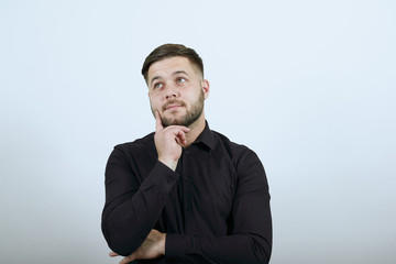 Young Bearded Dark Haired Man In Black Stylish Shirt On White Background, Serious Male Holds Forefinger To His Face And Thinks. The Concept Of Smart People.