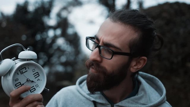 Bearded young man in glasses with long hair holds in his hand the alarm clock and a light bulb. Light bulb represents idea. Idea time concept.
