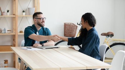 Happy businessman shaking hands with diverse woman job seeker. Smiling successful manager making deal with african american partner near laptop. Professional employee congratulations applicant.