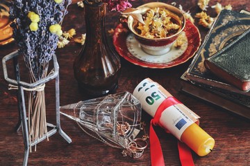 Money spell on wiccan witch altar - a red ribbon tied around a yellow candle with money banknotes. Slightly blurred background with focus on the money and vintage golden jewelry on dark wooden table