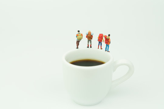 Miniature people.Male and femel tourists carrying a backpack behind. Standing top of the edge of the white coffee cup and black coffee inside.white background