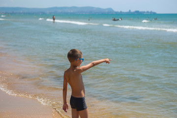 A child in sunglasses and swimming trunks looks at the ocean. A young boy points at the sea.