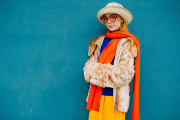 Unusual stylish teenager in a red sunglasses went for a walk in a blue sweater, yellow skirt and a jacket with a long orange scarf and white hat