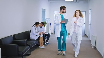 In front of the camera young nurse man and his colleague female doctor walking through the hospital corridor and discussing some diagnostic of the patient background other hospital doctor and nurses