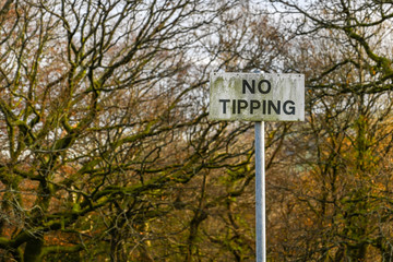 White "No tipping" sign covered in green algae isolated against a background of trees without leaves