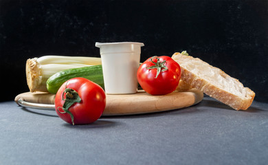 Slices of white beautiful airy white bread, yogurt, fresh vegetables - cucumbers, tomatoes and celery, all on a round wooden cutting board on a black background close-up, side view. Place for text.