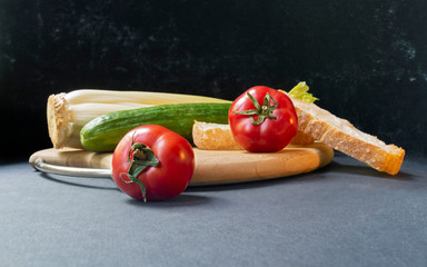 Two slices of white beautiful airy white bread, fresh vegetables - cucumbers, tomatoes and celery, all on a round wooden cutting board on a black background close-up, side view.