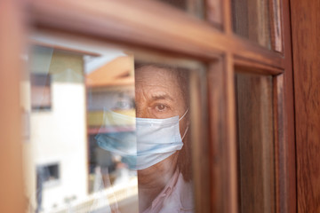 Ritratto di anziana che guarda dalla finestra di casa sua con l mascherina e aria triste
