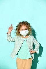Little girl in casual clothes. She is in medical mask, pointing at something, posing on turquoise background. Coronavirus concept