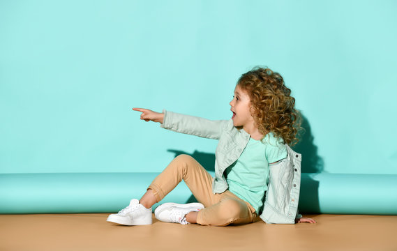 Nice little girl sits on the floor pointing finger aside commanding someone what to do. Side view portrait on turquoise background