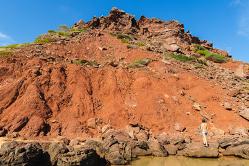 sensual unrecognizable woman in bikini looks at the mountain landscape and red sand on the beach el pilar in menorca (balearic islands, spain)