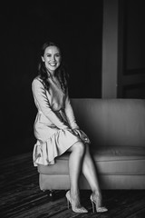 A beautiful young dark-skinned woman with dark curly hair and brown eyes in a gold dress sits on a mustard sofa in the loft room and smiles. Soft selective focus. Black and white photo.