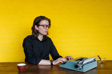 Serious young man writer with eyeglasses, wearing in black shirt, seated on a table, working on a typewriter, over yellow wall.