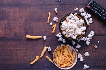 Top view of a bowl of popcorn, chips and tv remote on wooden background 