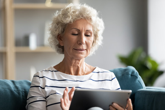 Head shot smiling old senior retired woman sitting on sofa, using computer tablet alone in living room. Happy elderly mature lady web surfing information, chatting with friends in social network.