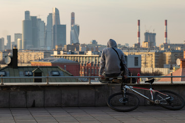 A man with a bicycle with his back to the camera admires the city in the setting sun.