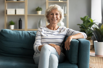 Obraz premium Front view beautiful grey haired sincere smiling mature senior woman leaning at sofa, enjoying leisure time in living room. Happy hoary middle aged pretty grandmother relaxing on couch alone at home.