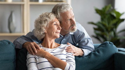 Dreamy pleasant middle aged married couple cuddling on sofa, looking away. Happy calm senior...