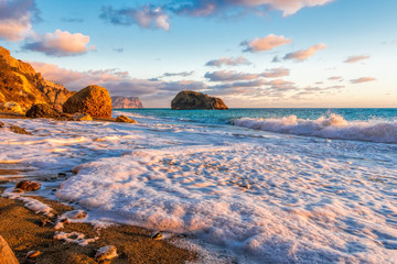 Bright sea sunset. The waves crash into the rock, lit by the warm sunset, sand and pebbles,...