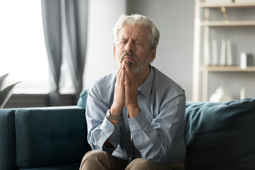 Stressed faithful older middle aged man praying begging God asking for help sitting on sofa with...