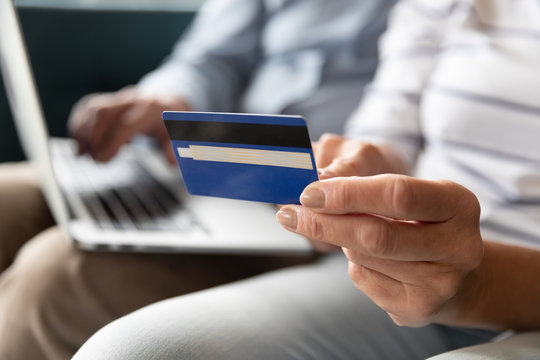 Close up elderly retired woman holding banking card in hand, paying utilities online or making purchases on computer in internet store together with husband, staying at home, easy technology usage.