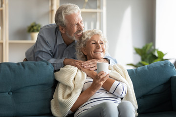Happy loving mature retired man taking care of smiling middle aged wife, covering her with plaid,...