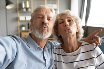 Web camera view childish middle aged married family couple having fun, taking kiss selfie photo at home. Playful older mature hoary man cuddling happy retired wife, recording funny video indoors.