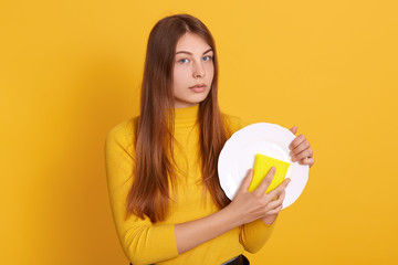 Image of beautiful young Caucasian woman looking directly at camera washing dishes in kitchen, posing against yellow wall, holding white plate and sponge in hands, dresses casually. Household chores.