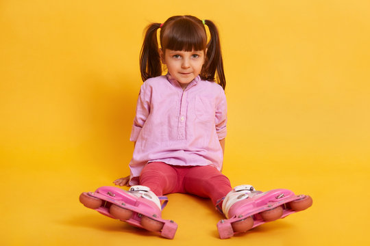 Image of adorable dark haired female child sitting on floor and looking directly at camera with serious facial expression, wearing tunic, leggins and rolling skates, having two ponytails. Childhood.