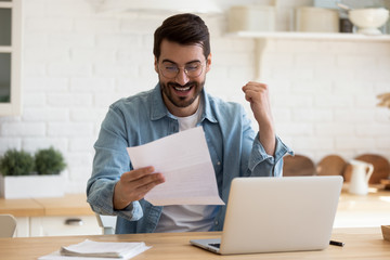 Excited millennial man in glasses sit at table in home kitchen feel euphoric reading good news in...