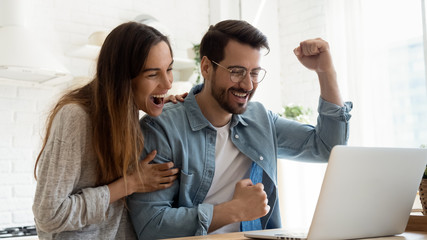 Overjoyed millennial couple sit at desk look at laptop screen feel euphoric with online win,...