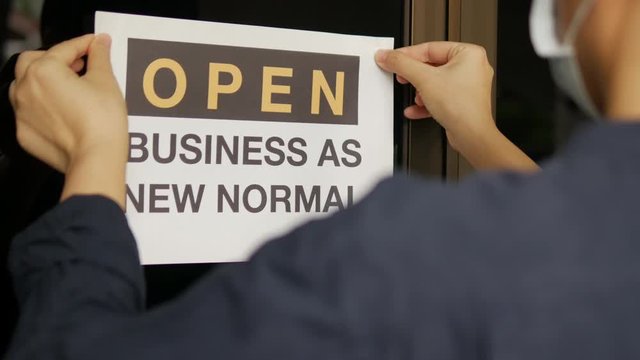 Reopening for business adapt to new normal in the novel Coronavirus COVID-19 pandemic. Rear view of business owner wearing medical mask placing open sign &ldquo;OPEN BUSINESS AS NEW NORMAL&rdquo; on front door.