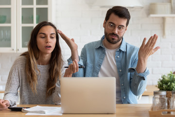 Mad young husband and wife sit at desk in kitchen feel frustrated with slow Internet connection on computer, angry millennial couple manage household finances get operational problem on laptop