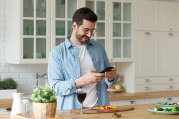 Smiling Caucasian young man use cellphone gadget texting messaging while preparing food in kitchen, happy millennial male read recipe on smartphone get ready for home party or celebration