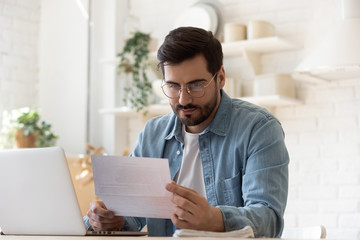 Pensive millennial man in glasses sit at table work on laptop read letter correspondence thinking,...