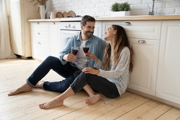 Happy millennial couple sit relax on wooden floor in kitchen drinking red wine talking chatting,...