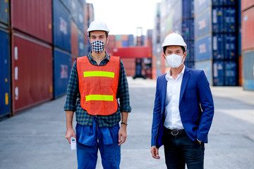 Technician or engineer workers stand in front of shipping container with one hold infrared thermometer and manager also wear face mask in the procedure to prevent virus infection in workplace area.