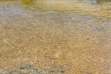 Yellow-orange pebbles can be seen through the clear clear water on a sunny beach. Abstract. Background.