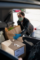 Young female in protective mask and gloves putting paperbags into car trunk