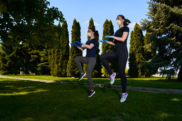 Obraz premium Two girls do sports exercises in protective masks in the open air in park. Social distancing. Alternative fitness workouts during epidemic and quarantine. Covid 19 pandemic. Coronavirus