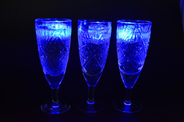 Crystal champagne glasses with carvings in the form of snowflakes are located on a black matte background. Bright multicolor doidic glowing ice cubes float in the drink.