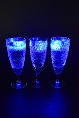 Crystal champagne glasses with carvings in the form of snowflakes are located on a black matte background. Bright multicolor doidic glowing ice cubes float in the drink.