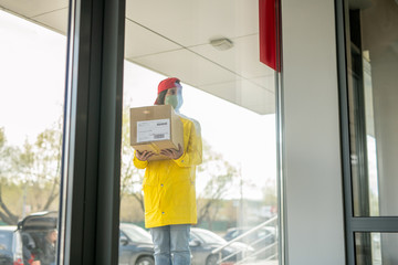 Young woman in workwear and protective mask holding packed box with order