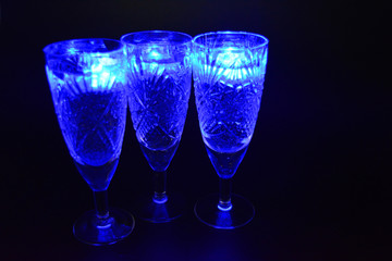 Crystal champagne glasses with carvings in the form of snowflakes are located on a black matte background. Bright multicolor doidic glowing ice cubes float in the drink.