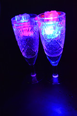 Crystal champagne glasses with carvings in the form of snowflakes are located on a black matte background. Bright multicolor doidic glowing ice cubes float in the drink.