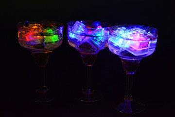 Three open champagne glasses with luminous multicolor ice cubes are located on a black matte background.