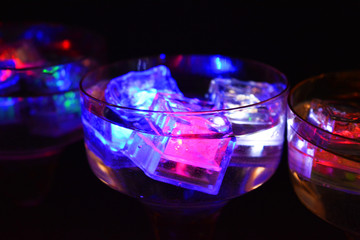 Three open champagne glasses with luminous multicolor ice cubes are located on a black matte background.