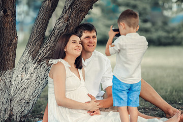 A young photographer. The child takes pictures of his mother and father on a summer day under a tree. Funny parents smile at the camera.